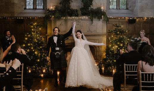 a cheering bride and groom standing in front of a large stone fireplace that's illuminated by fairy lights and has Christmas trees either side.