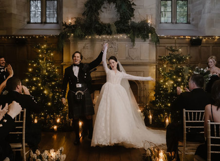 a cheering bride and groom standing in front of a large stone fireplace that's illuminated by fairy lights and has Christmas trees either side.