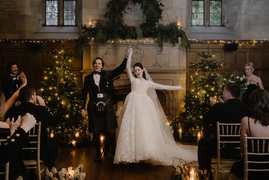 a cheering bride and groom standing in front of a large stone fireplace that's illuminated by fairy lights and has Christmas trees either side.