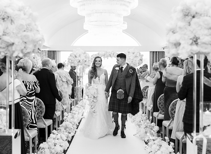 a bride and groom walking up an aisle holding hands with rows of seated guests on either side. There is a white carpet lined with flower arrangements on either side and tall white rose flower arrangements in foreground