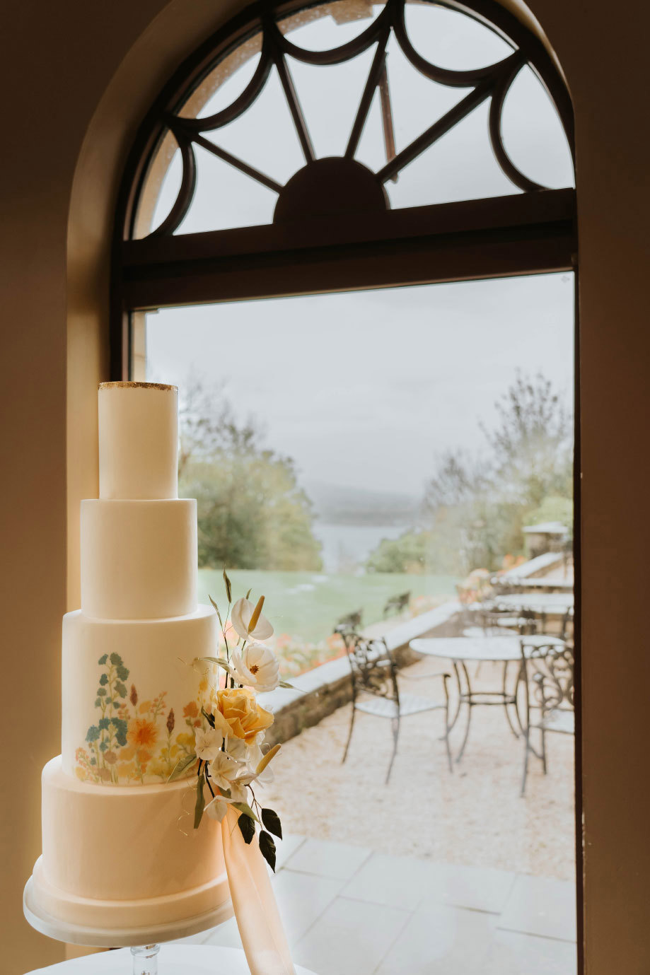 A four tier wedding cake with a floral desing sitting next to a window looking onto a courtyard 