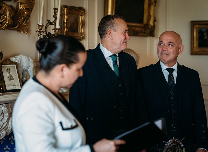 two grooms look at one another emotionally during a wedding ceremony as a person reads in front of them