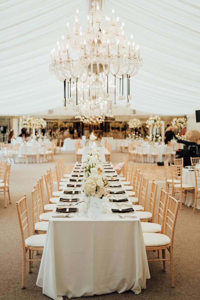 A long table underneath a glass chandelier inside a marquee with other round tables in the background 