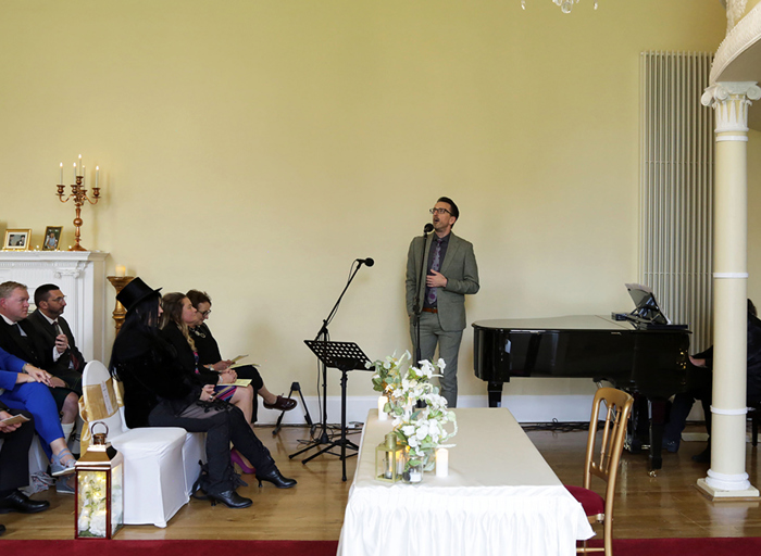 A man stands at a microphone singing next to a black piano 