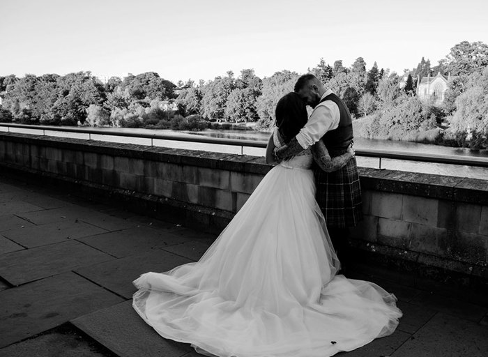 Black and white photo of a bride in a ballgown wedding dress and a man in a kilt on a stone bridge