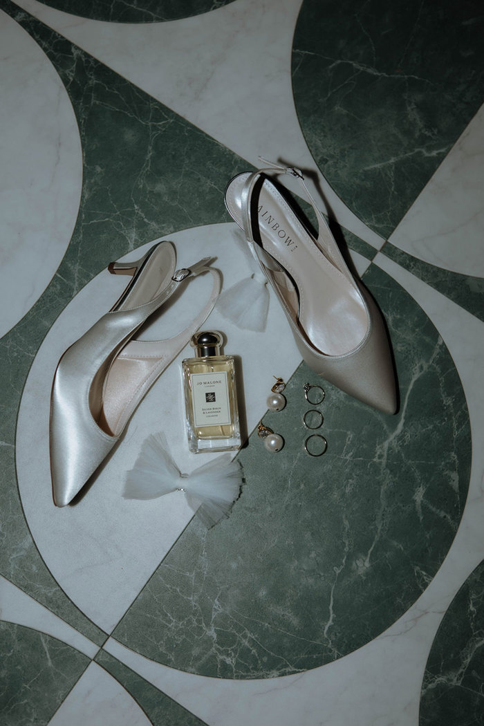 white satin high heels lying on a tile floor next to a bottle of Jo Malone perfume, tulle bows, pearl earrings and silver rings