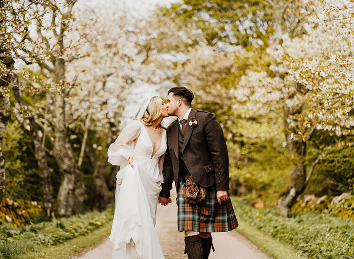 a bride and groom kiss while walking along a path surrounded by trees with white blossom either side of path