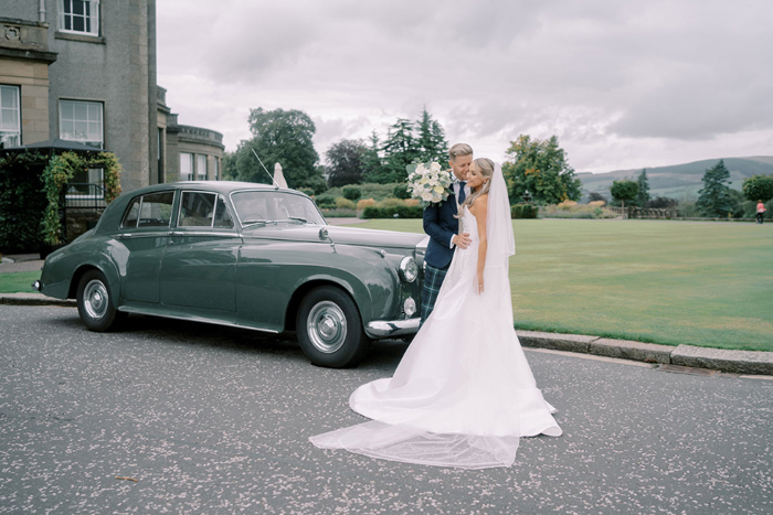 Bride and groom pose in front of wedding car