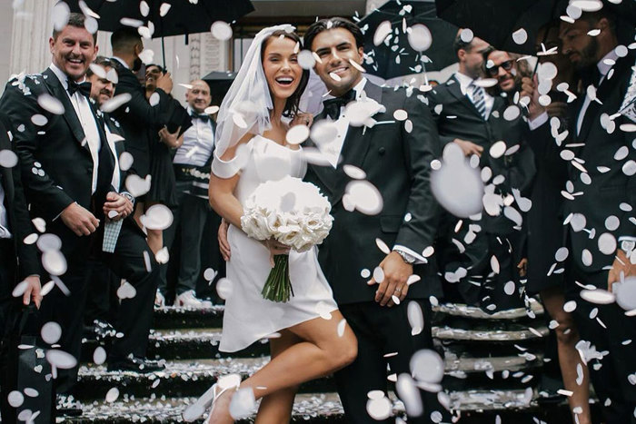 A bride wearing a short dress and veil and a groom wearing a black tuxedo being showered in white confetti