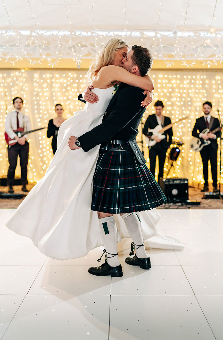 bride and groom kissing on a white dance floor. The groom is lifting the bride up and they are embracing. There is a band in the background and an illuminated wall of fairy lights