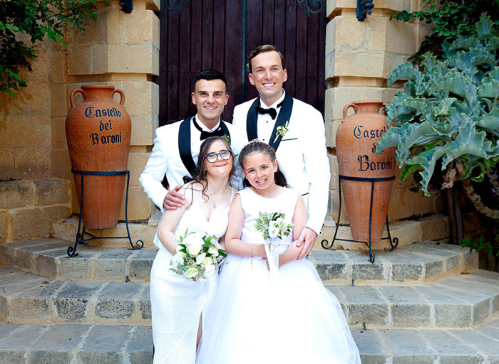 two grooms wearing smart white suit jackets and bow ties posing with two flower girls wearing white dresses. They are standing in front of an old stone building and steps with large terracotta urns either side