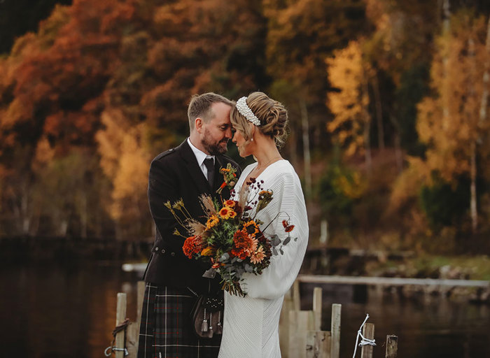 a bride wearing a chunky pearl headband and holding an autumn-toned dried flower bouquet stands head to head with a groom wearing a kilt. There are autumn trees of gold and russet in the background