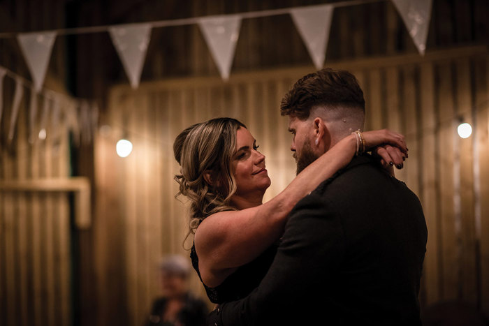 A Bride And Groom Embrace In A Wooden Building With White Bunting Hanging Above And In Background