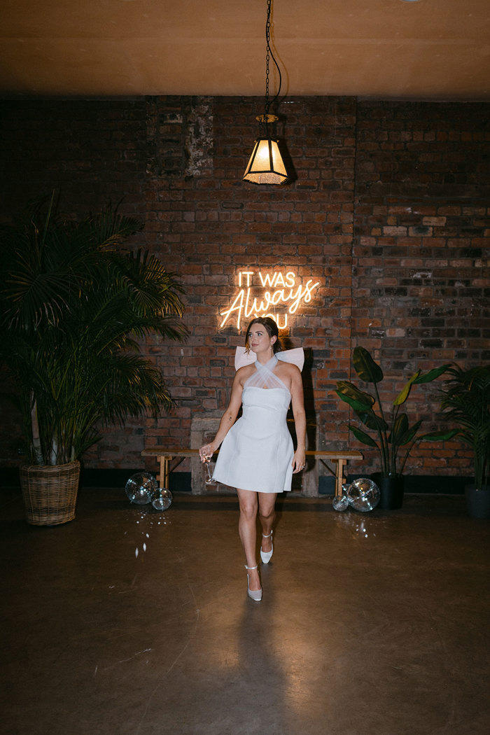 a bride wearing a short dress walking in front of a brick wall and neon sign 