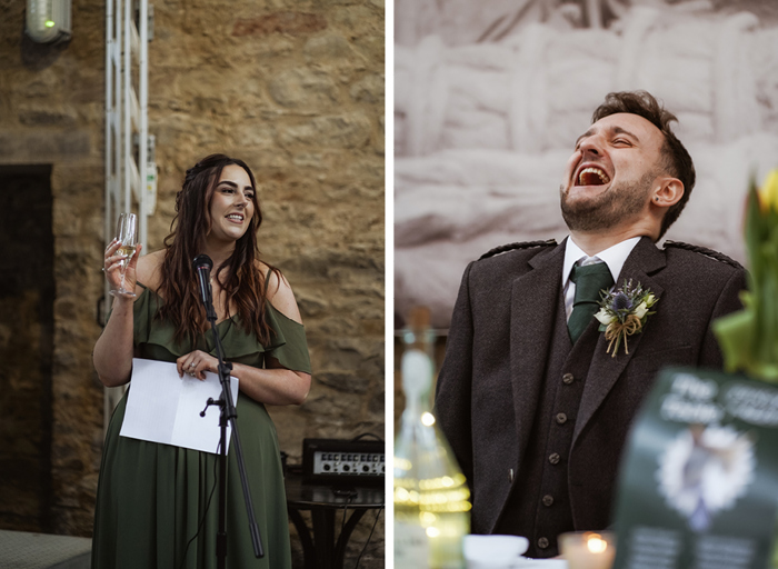 a person wearing a green dress holding a champagne flute and a piece of paper standing in front of a microphone on left. A person seated at a table wearing a grey tweed jacket, tie and buttonhole throwing their head back while laughing on right