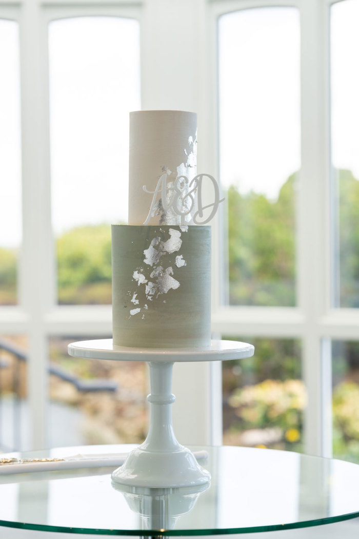 an elegant tall wedding cake with silver foil detail sitting on a white cake stand in front of a window