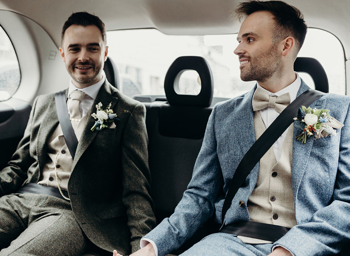 two grooms in seated in the back of a taxi holding hands. One is wearing a dark green tweed suit and cream tie and waistcoat and the other is wearing a blue tweed suit and beige bow tie and waistcoat