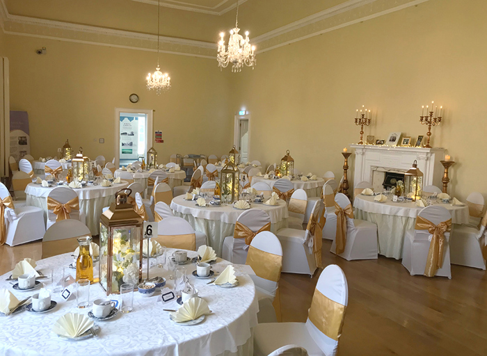 A room with several round tables covered in white table cloths which have chairs around them in white chair covers with gold ribbon tied around them. The tables are set for an afternoon tea