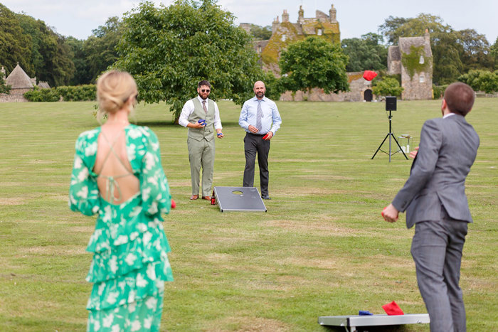 four people stand across the grass from each other playing cornhole 