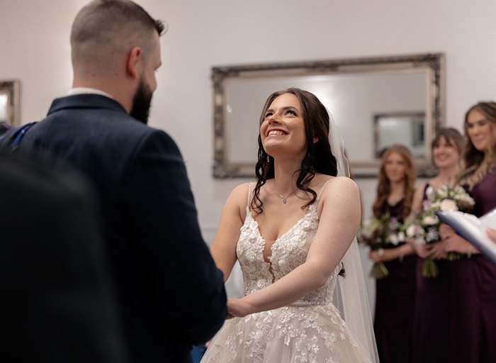 a bride wearing a floral detail ivory dress. She is throwing her head back laughing while holding the hands of a groom in a kilt who stands in front of her. Bridesmaids wearing purple dresses are in the background