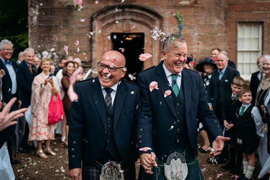 two grooms wearing kilts walk hand in hand while being showered by guests in flower confetti outside Brodick Castle