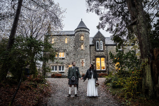 a Scottish wedding day couple holding hands outside a small castle-like building