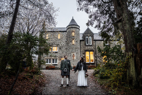 a Scottish wedding day couple holding hands outside a small castle-like building