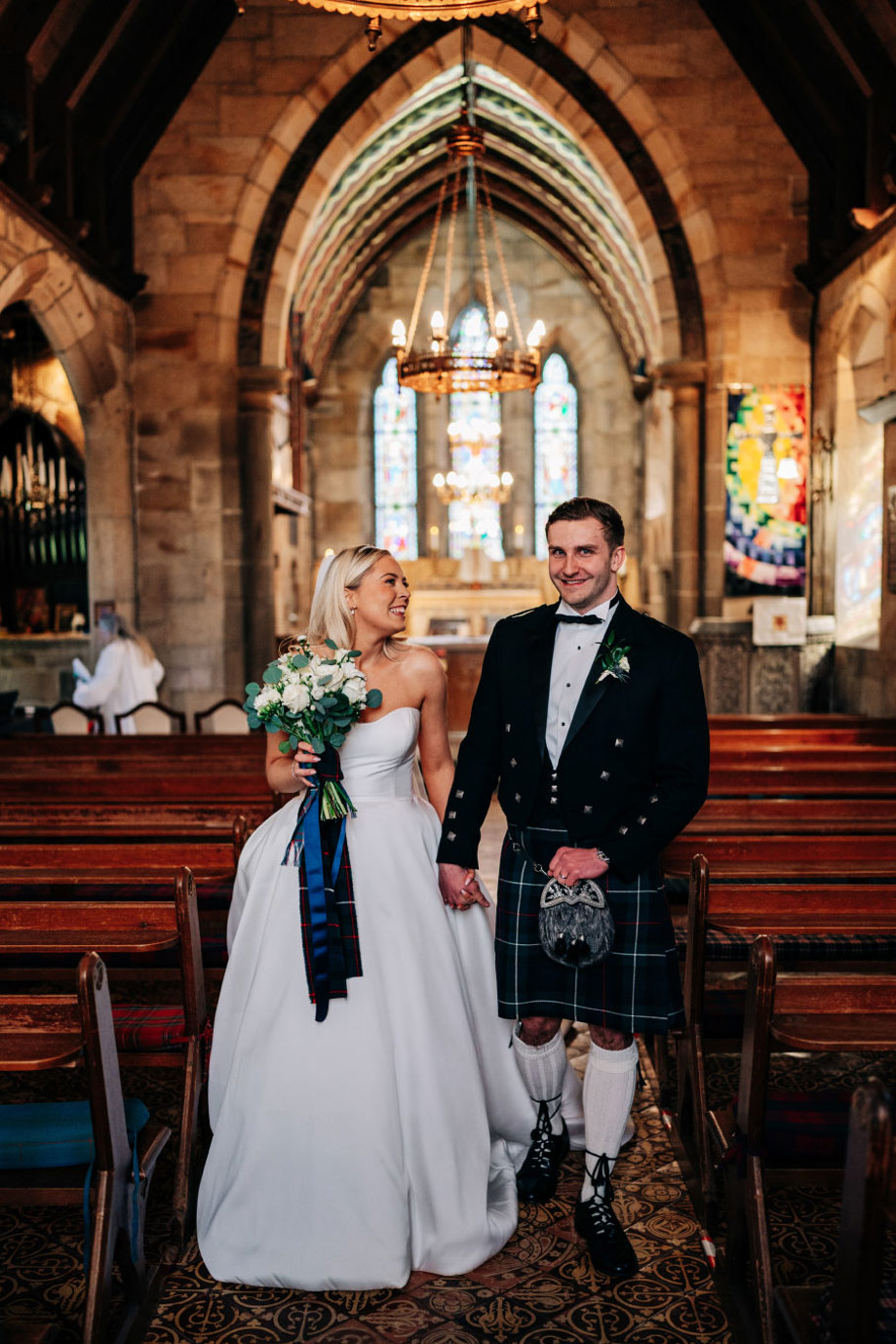 a smiling bride and groom holding hands walking up an aisle in a church