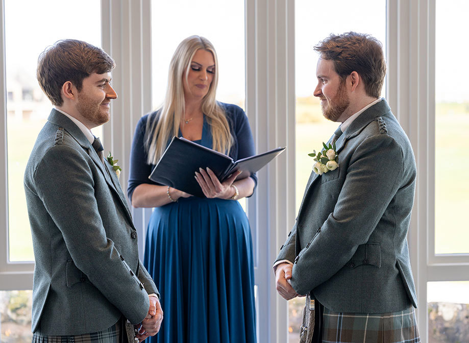 two grooms wearing kilts face each other and smile as a celebrant behind them reads from a book