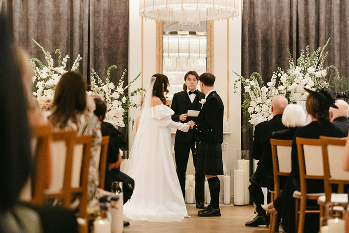 brunette bride in long veil and wedding dress with puff sleeves and groom in full dark tartan kilt outfit hold both of each other's hands in front of them at the top of the aisle as another man in a tux stands reading from a piece of paper