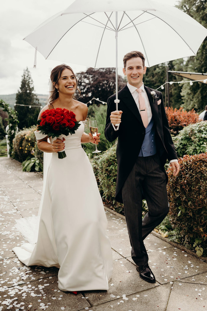 A groom holding an umbrella and bride holding a bouquet of red roses walking along a path in a garden setting