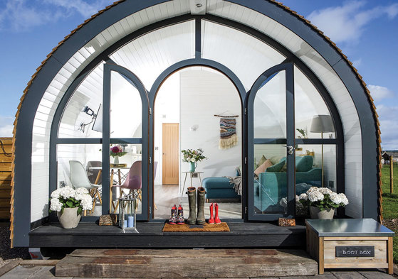 the front view of a pod with windows making up the entire front, including the door, has several pairs of wellies sat in front and a living and dining room visible within