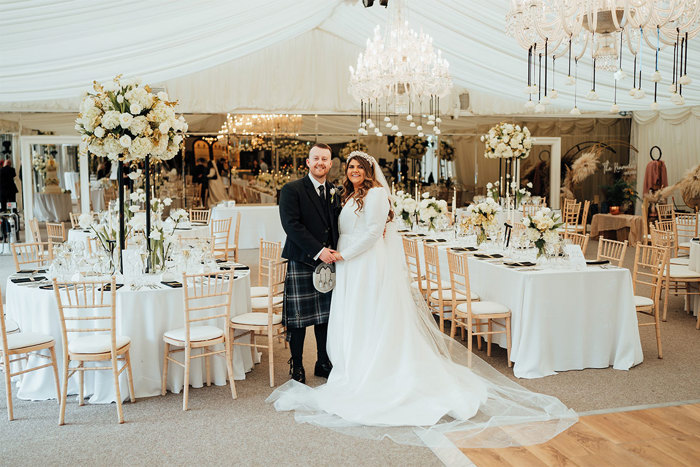 bride and groom standing in marquee surrounded by tables with flowers and chairs