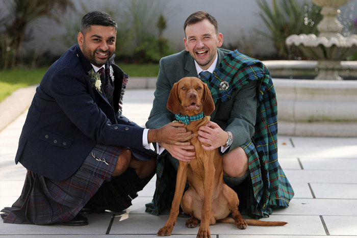 Two men in kilts crouch down next to a brown dog who is wearing a green tartan collar