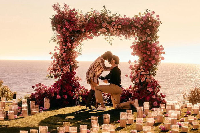 A proposal between a man and a woman in front of a pink flower arch at sunset overlooking the sea, the couple are surrounded by candles