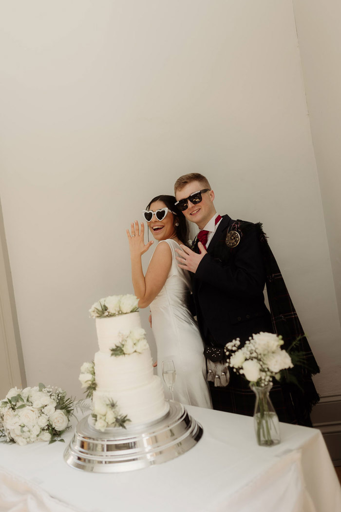 bride and groom pose with wedding rings next to cake at kilmardinny house in bearsden