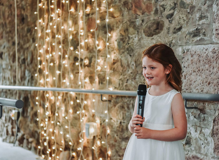 a young girl holding a microphone standing next to a stone wall with strings of fairylights in the background