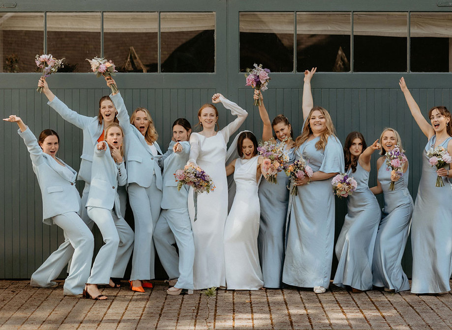 a fun line up of two brides and 10 bridesmaids wearing blue Whistles trouser suits and blue Six Stories posing against a dark teal painted wooden door