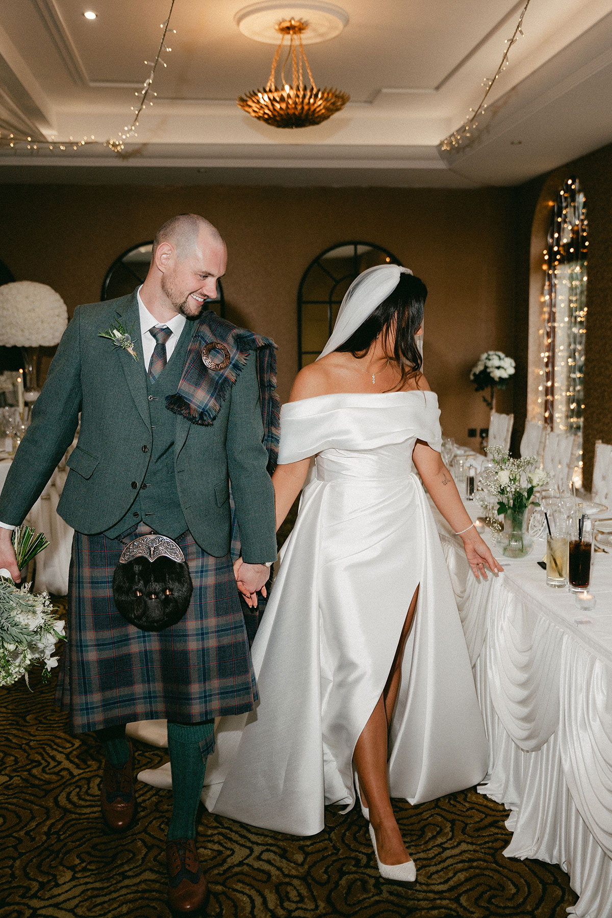 bride with long veil and off the shoulder wedding dress with a leg slit holds hands with groom in green, blue and purple tartan kilt outfit with green jacket admiring reception table displays