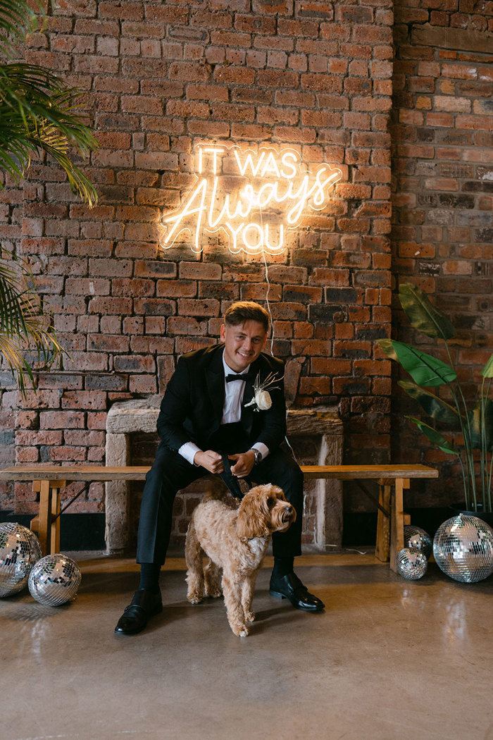 a dog and a man in a suit sitting on a bench in front of a brick wall below a neon sign at the Engine Works