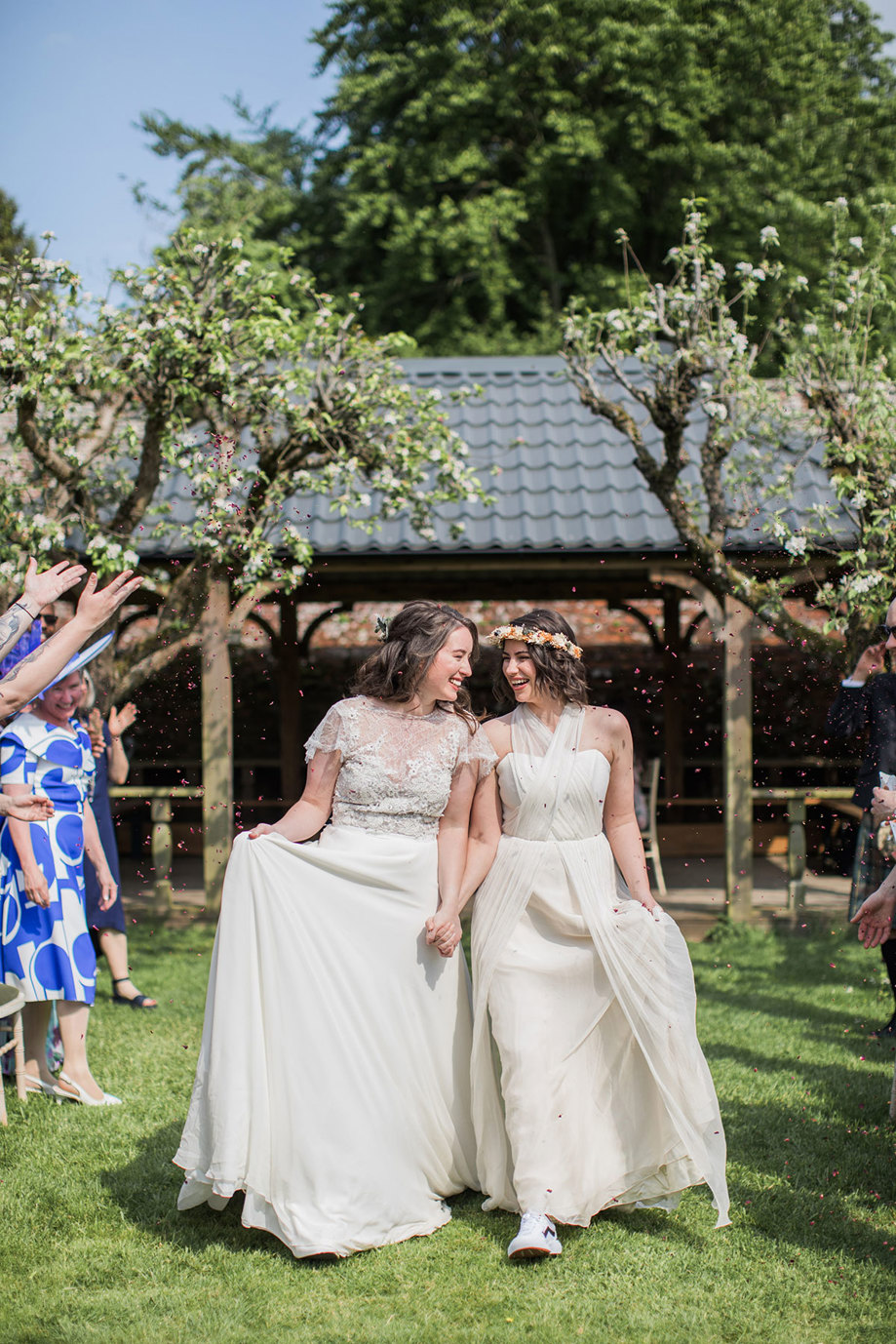 two elated brides walking hand-in-hand across grass. They are being showered in dark red confetti and there is a roofed wooden structure in the background.