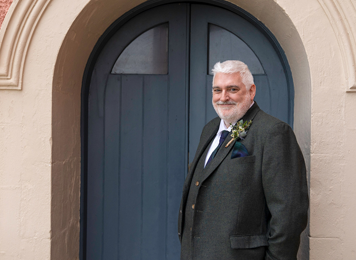 a man in a dark grey suit standing in front of a navy blue door with stone arched frame