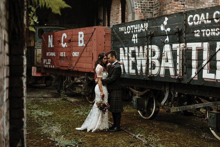 a bride and groom stand in an embrace in front of two train carriages