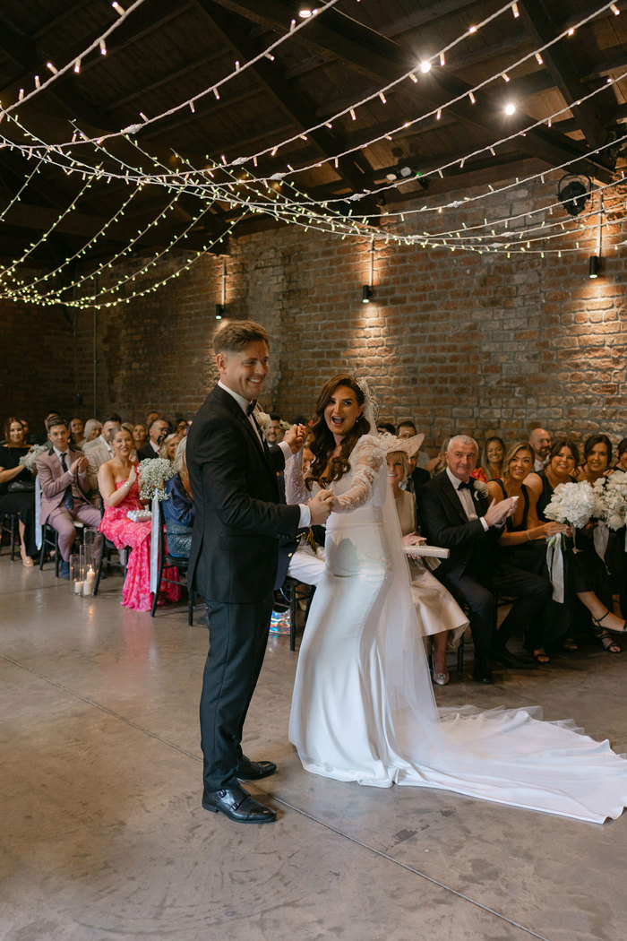 a bride and groom standing during a wedding ceremony at the Engine Works. There are rows of seated guests behind them watching on