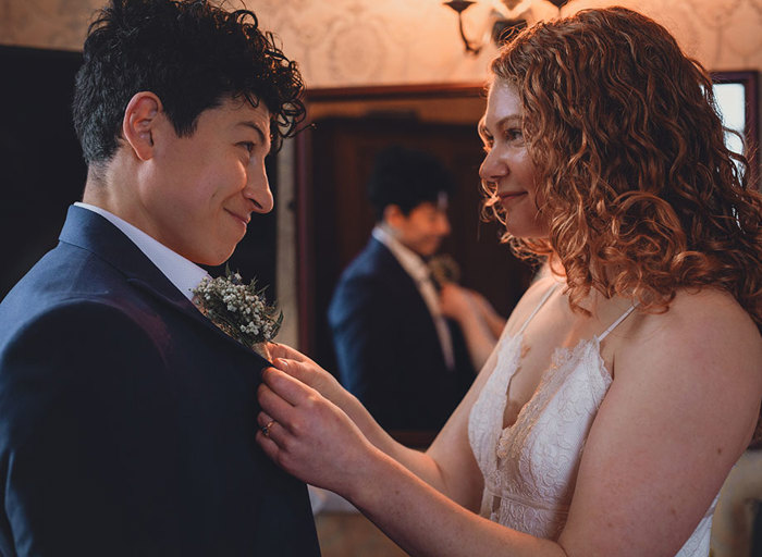 A bride in a suit and a bride in a white jumpsuit look into each other's eyes as one fixes the other's buttonhole