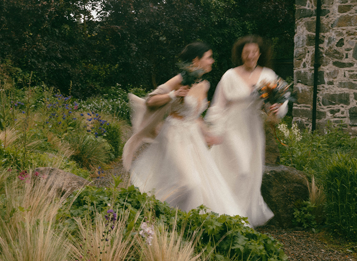 a blurred image of two brides holding hands waling through a flower filled garden with old stone building in right hand background