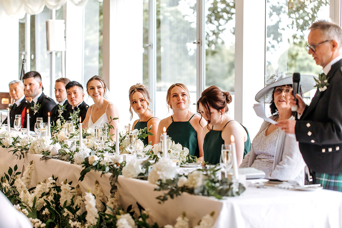 bride, groom and bridesmaids look towards father of the groom while sitting at long table with foliage at mar hall