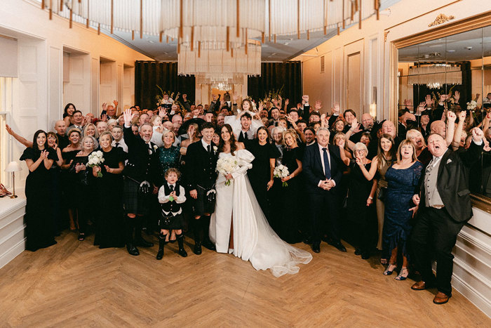 full room of a wedding group photo where brunette bride in high leg slit wedding dress with long puff sleeves and groom in full dark tartan kilt outfit stand in the centre with all others around them, holding their hands up in the air