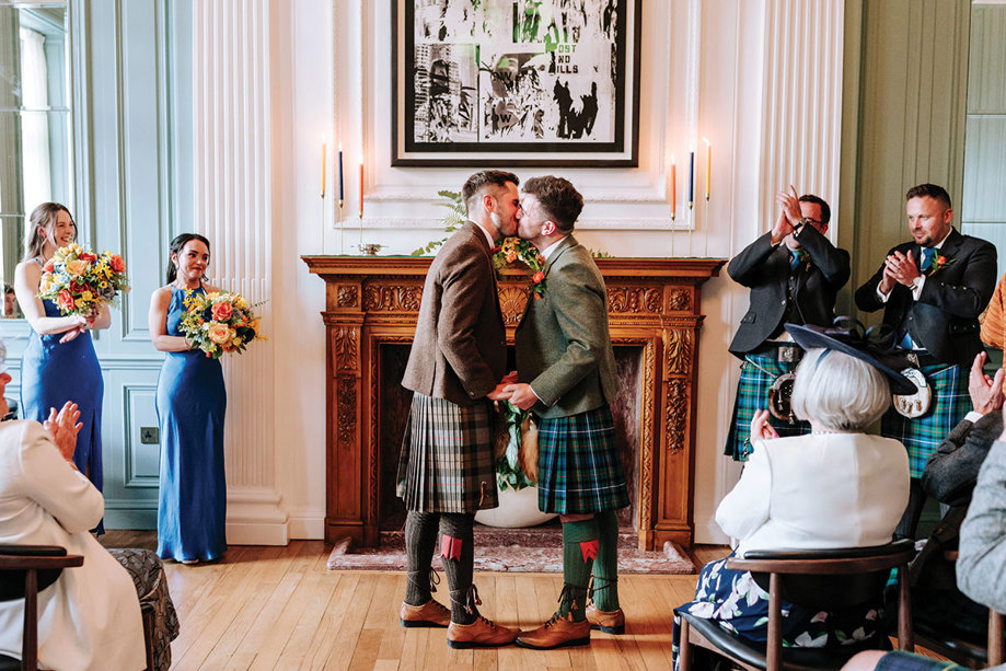 Two men kissing in the Games Room at Cheval Edinburgh Grand with people in the background.