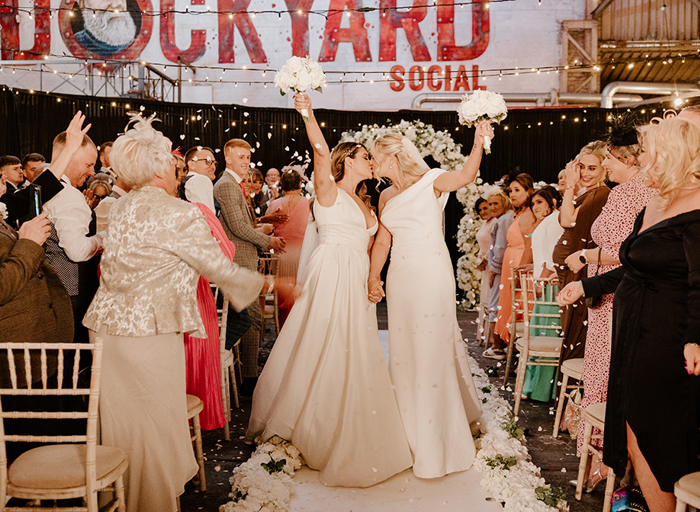 two brides kiss and hold their bouquets aloft while standing on the aisle after their Dockyard Social wedding ceremony. Guests cheer and throw confetti either side